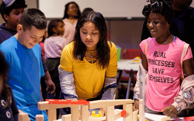 Three students, a boy in a blue shirt, a girl in a yellow shirt, and a girl in a pink shirt, stand over a marble rollercoaster that they created.