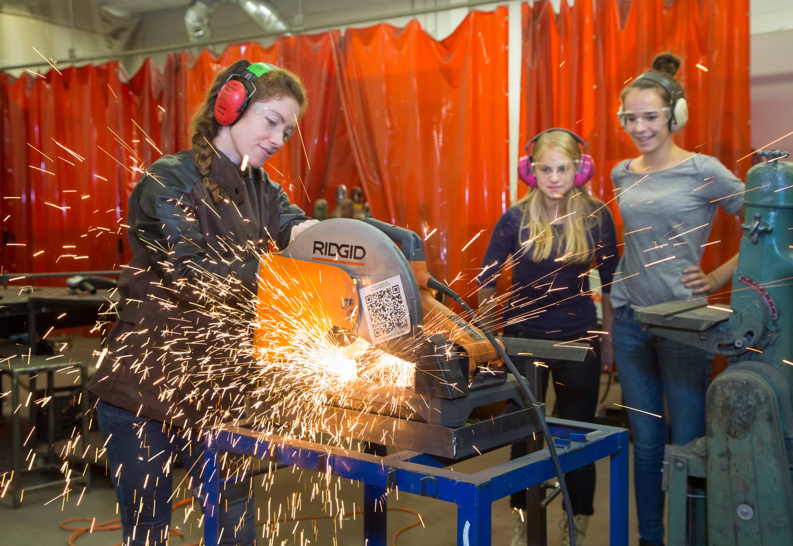 Woman engineer with 2 teenage girls creates sparks as she cuts metal.